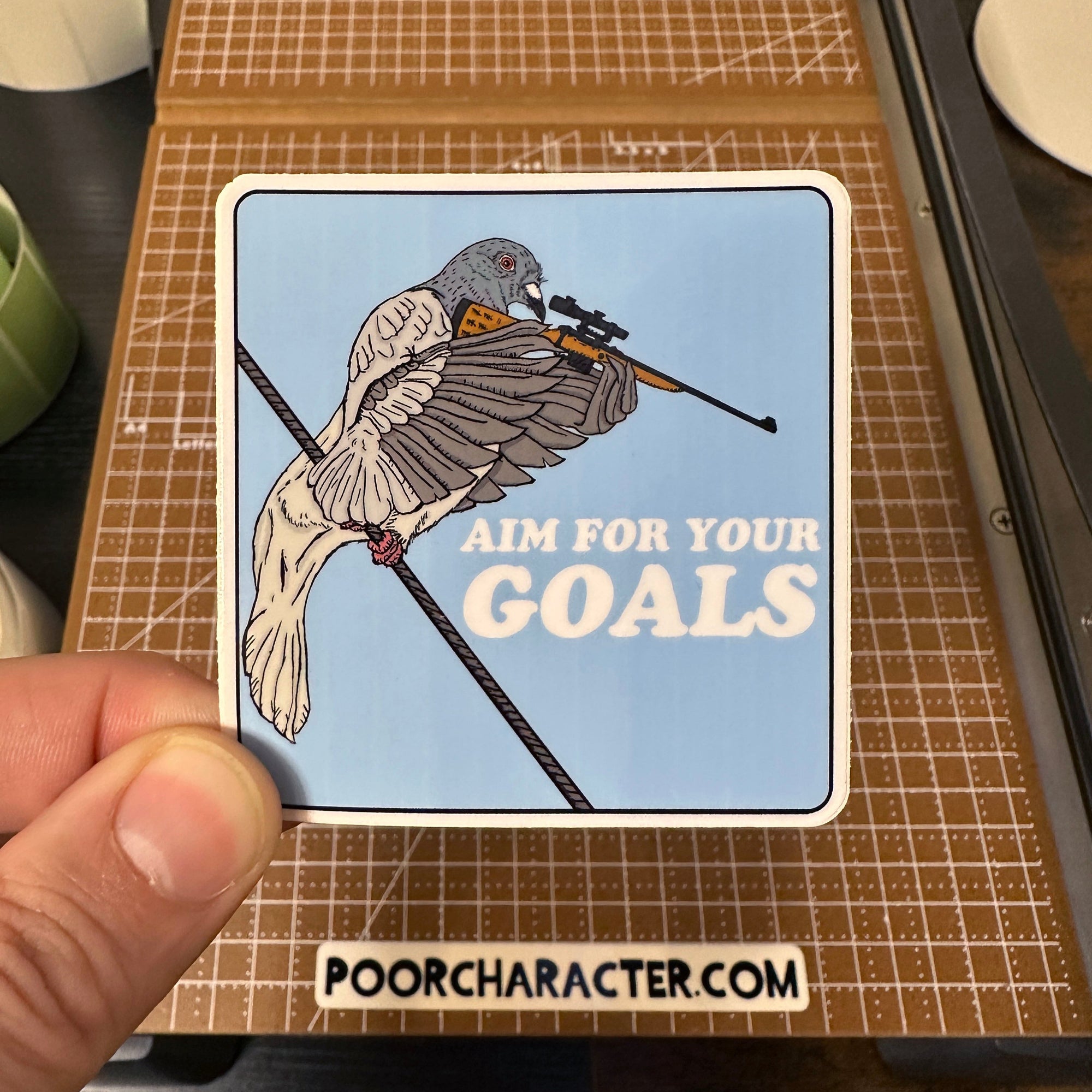 Sticker of A pigeon on a wire, aiming a sniper rifle with the words 'Aim For Your Goals' beside it