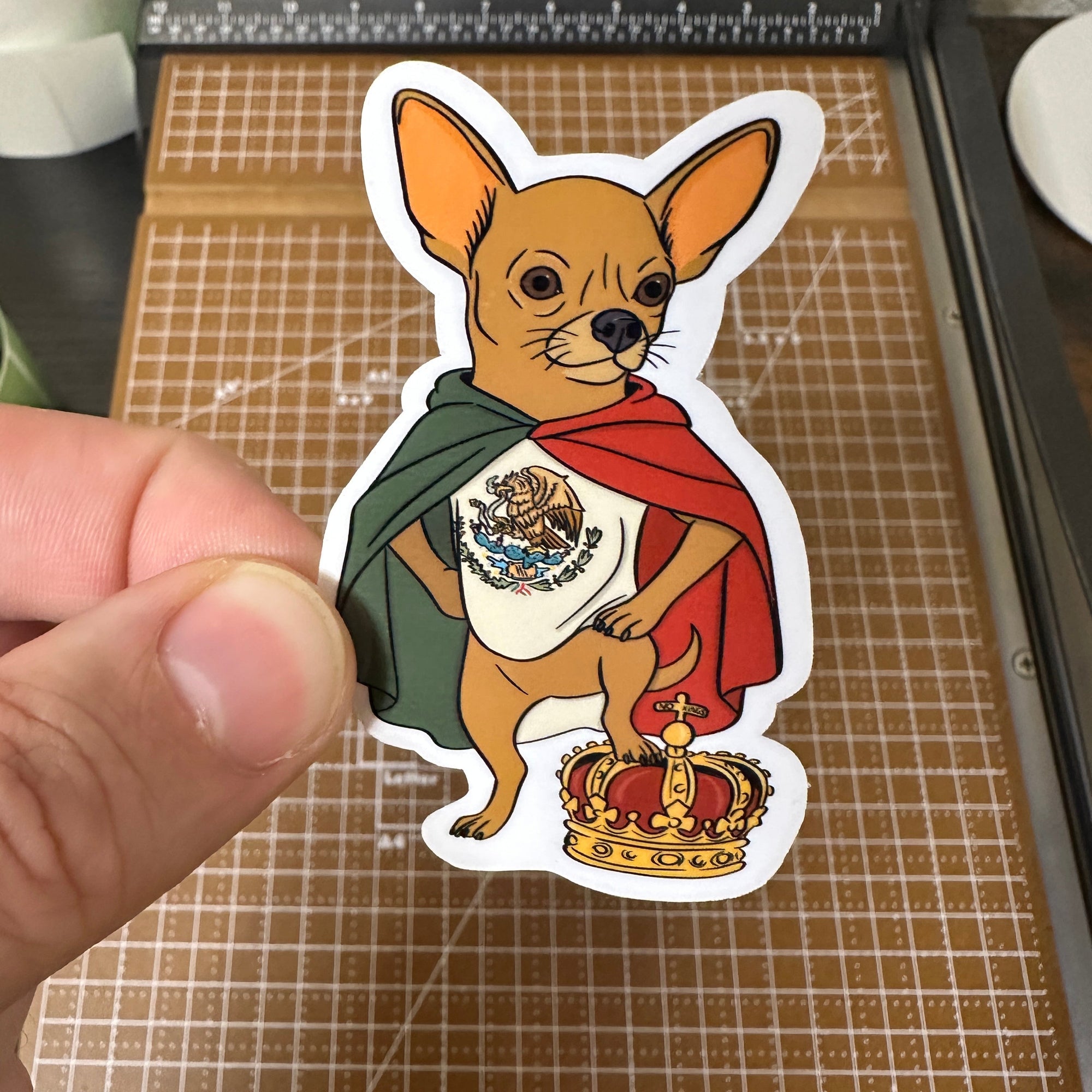 Photo of a Chihuahua sticker, draped in a Mexican flag standing with one foot on top of a crown 