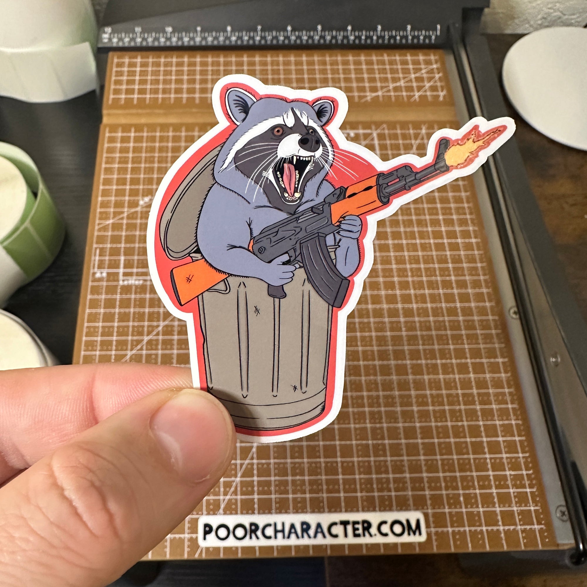 Sticker of a raccoon popping out of a trashcan while shooting an AK-47, drawn in a cartoonishly wild and rebellious style.