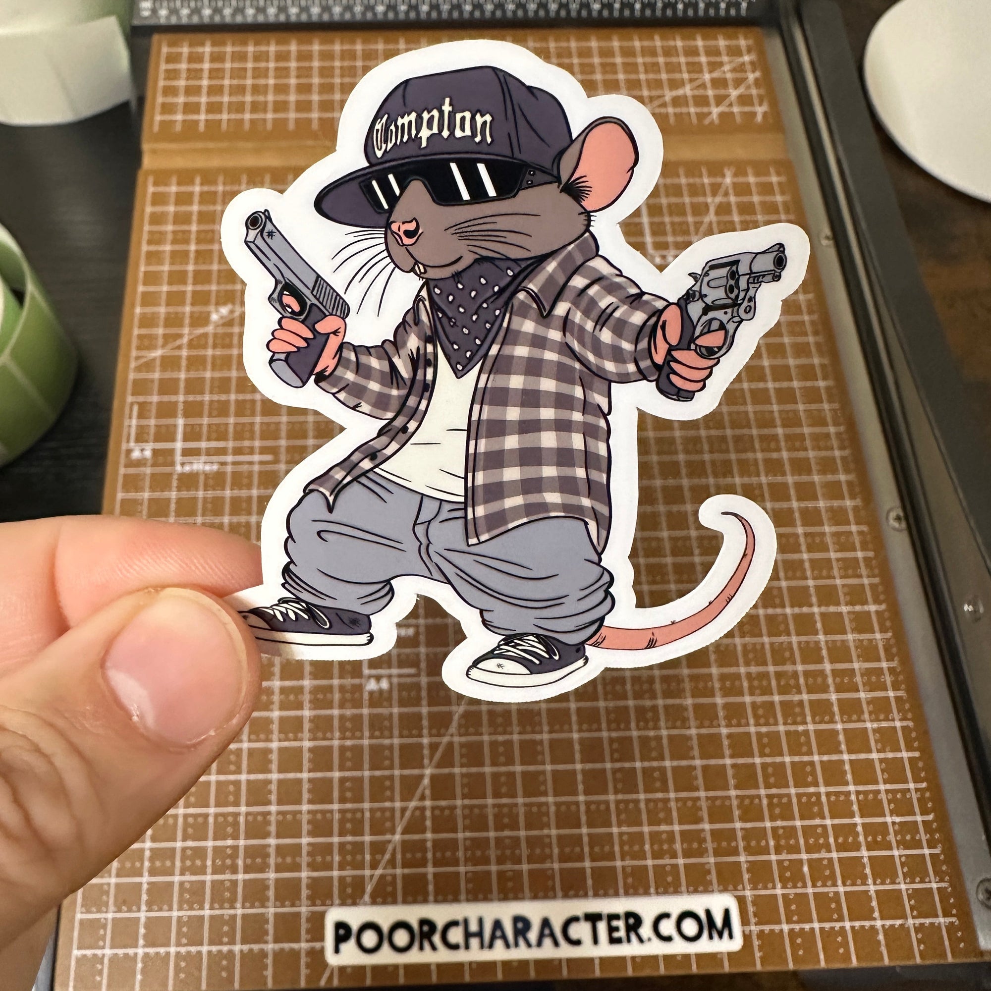 Sticker of a rat paying homage to Eazy E, holding two handguns, wearing a Compton hat and plaid shirt